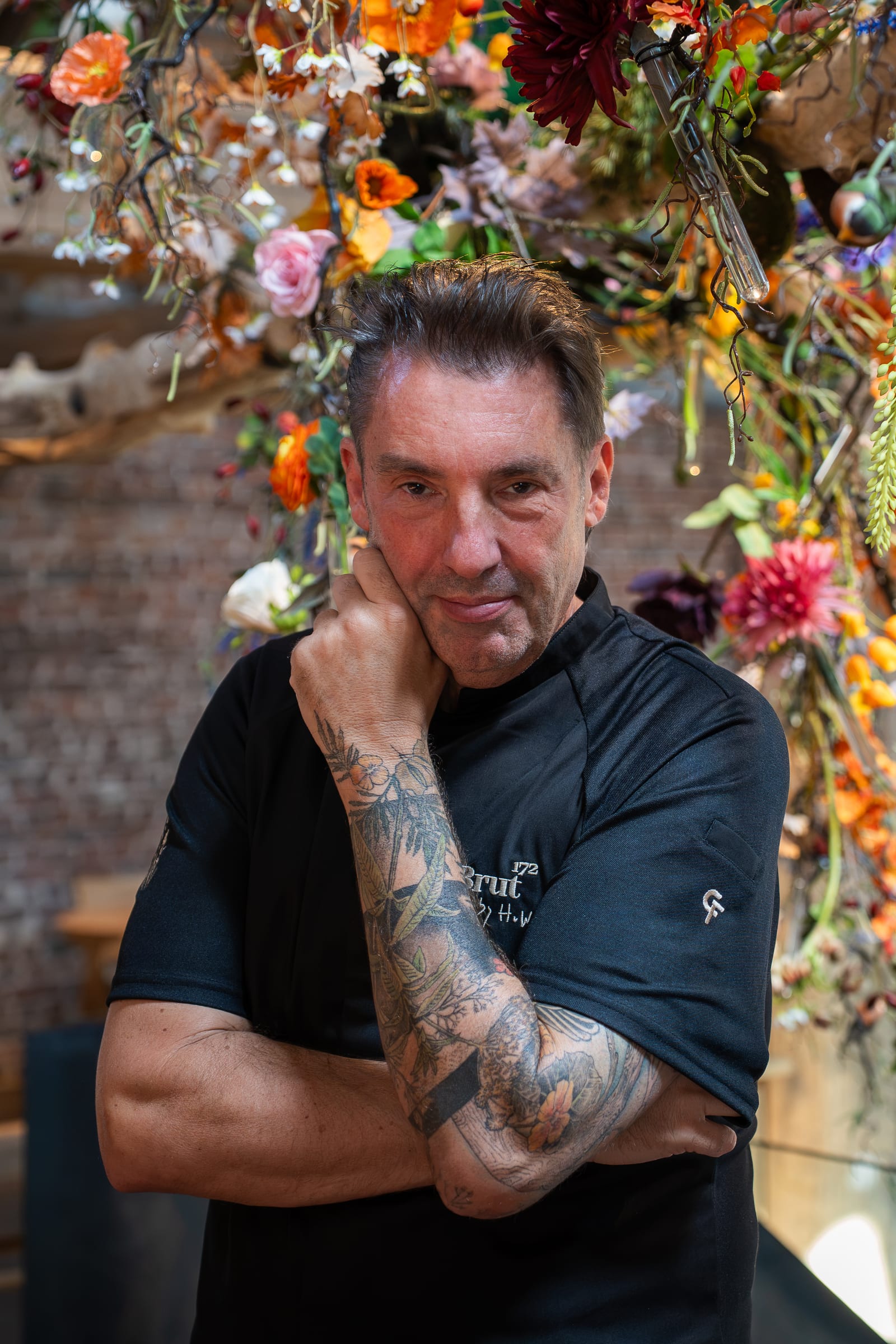 Hans van Wolde in een zwarte Cheffix-koksbuis, met zijn hand onder zijn kin, voor een plafond vol bloemen